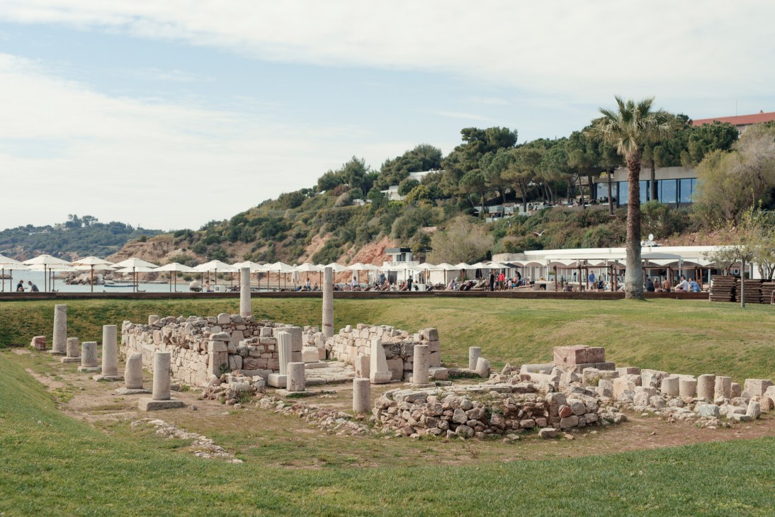 Ancient stone temple ruins with columns scattered across a grassy field overlooking a Mediterranean beach and coastline.
