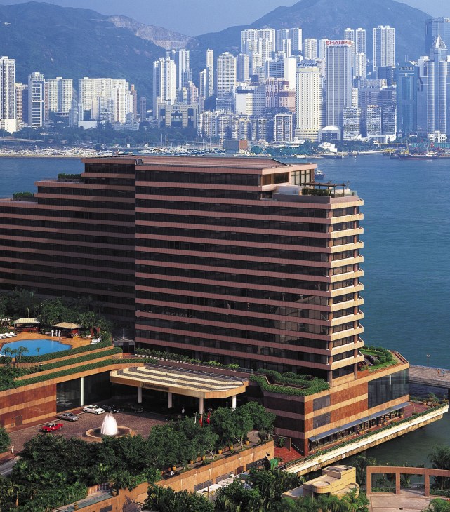 The Regent Hong Kong, a modernist multi-story hotel building with distinctive horizontal striping, overlooks Victoria Harbour with mountains and high-rise buildings in the background.