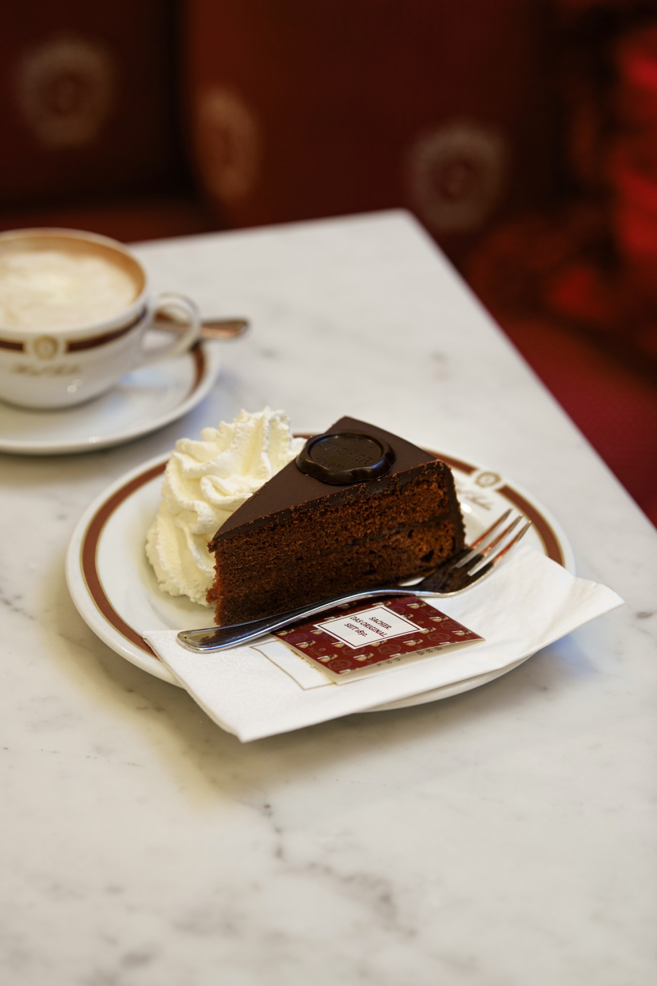 Slice of Original Sacher-Torte with whipped cream on white plate beside coffee cup