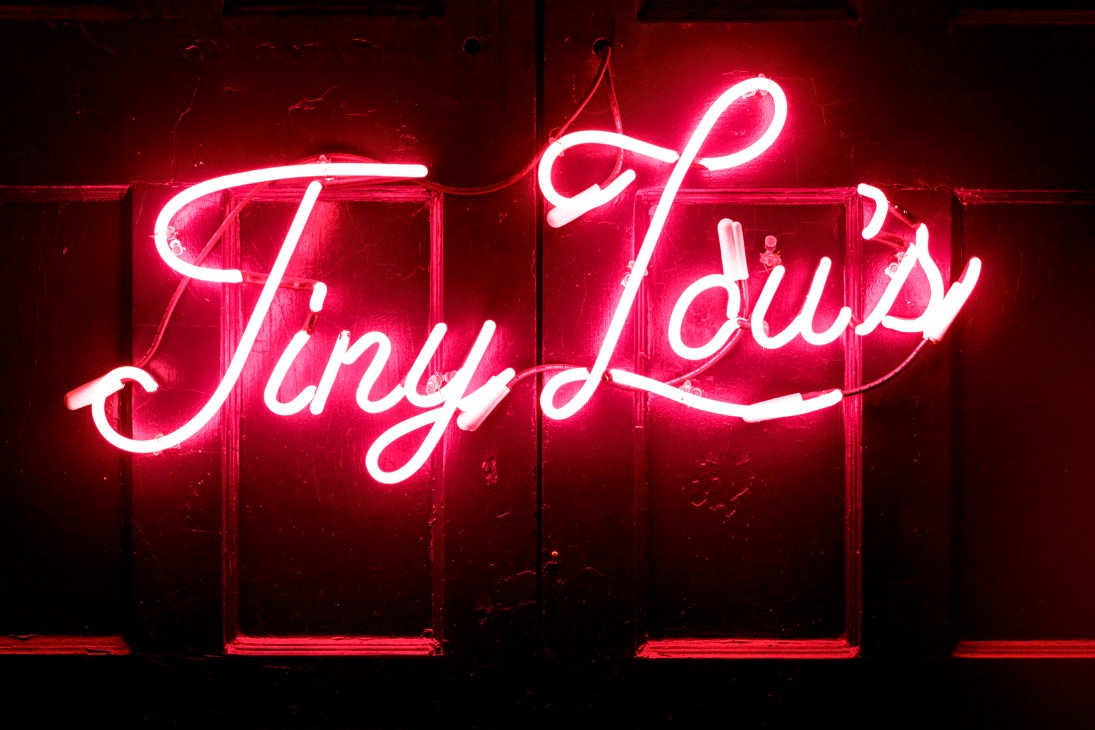 Pink neon cursive sign reading "Tiny Lou's" mounted on dark wall