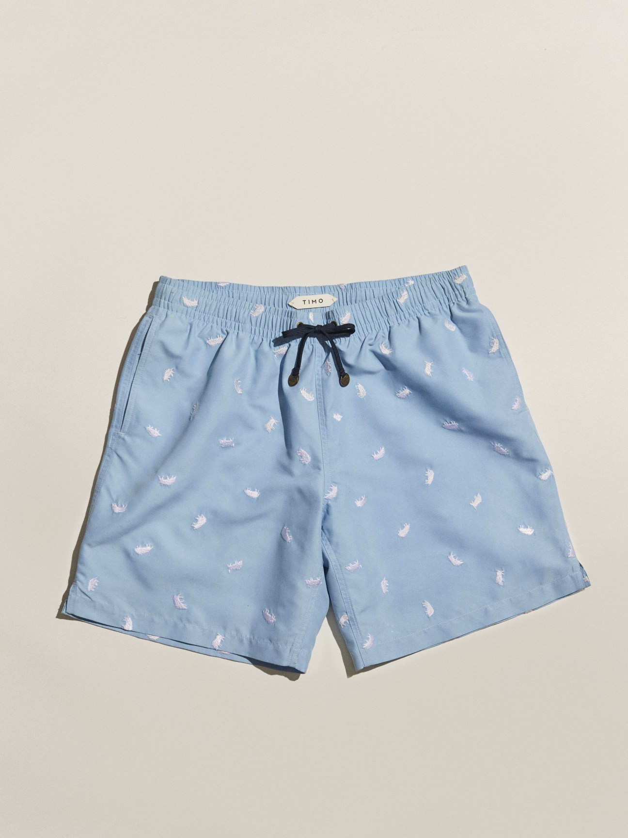 Light blue swimming shorts by Timo with white palm tree print and black drawstring on cream background.