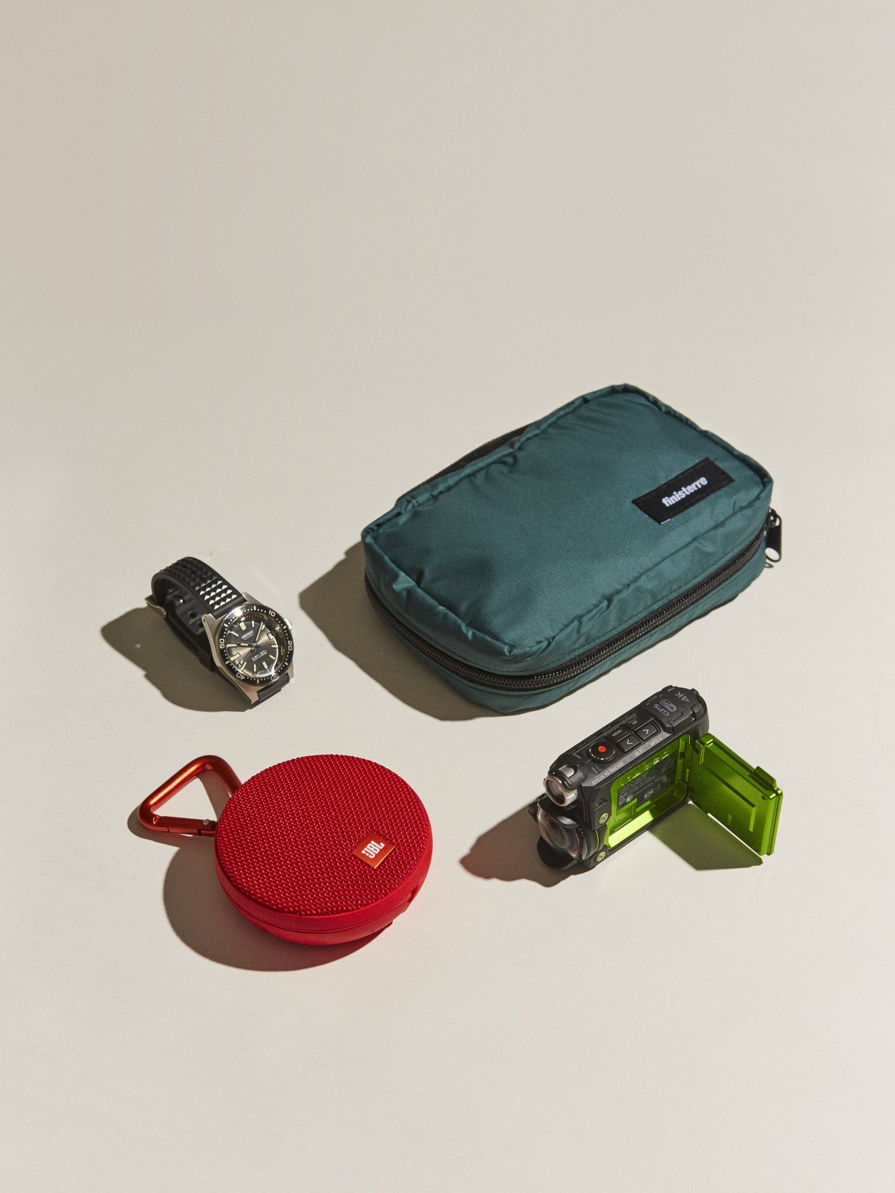 Watch by Seiko Prospex, pouch by Finisterre, camera by Olympus, and speaker by JBL arranged on neutral background.