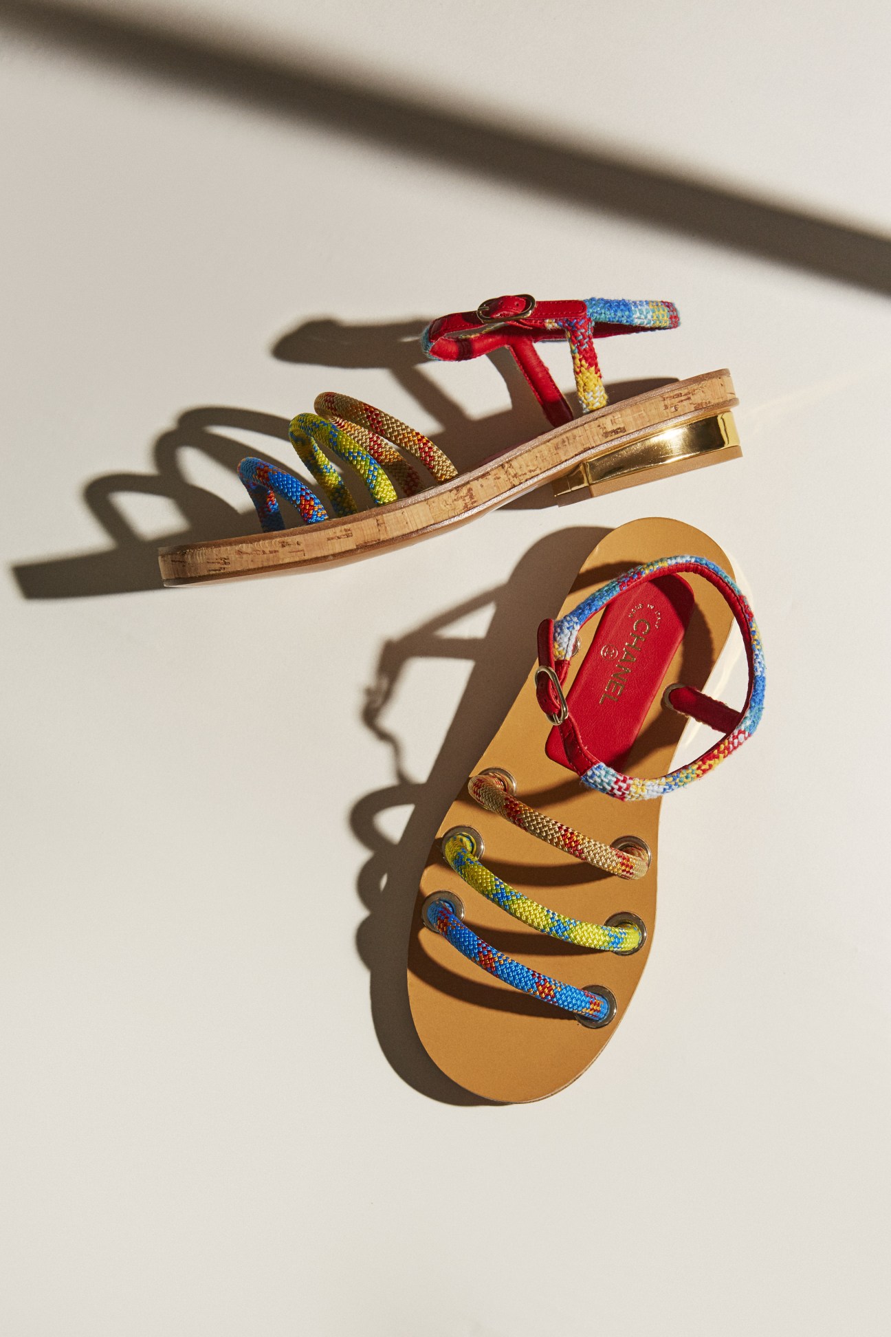 Chanel sandals with tan leather soles and colorful woven straps cast shadows on a white surface.