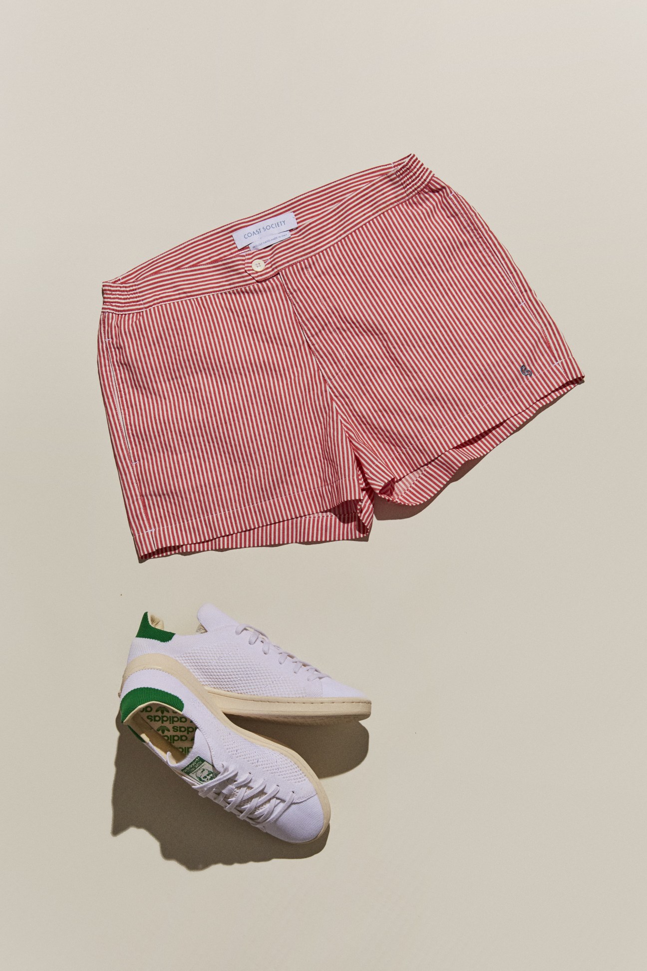 Red striped swimming shorts by Coast Society and white Adidas trainers for J.Crew on beige background.