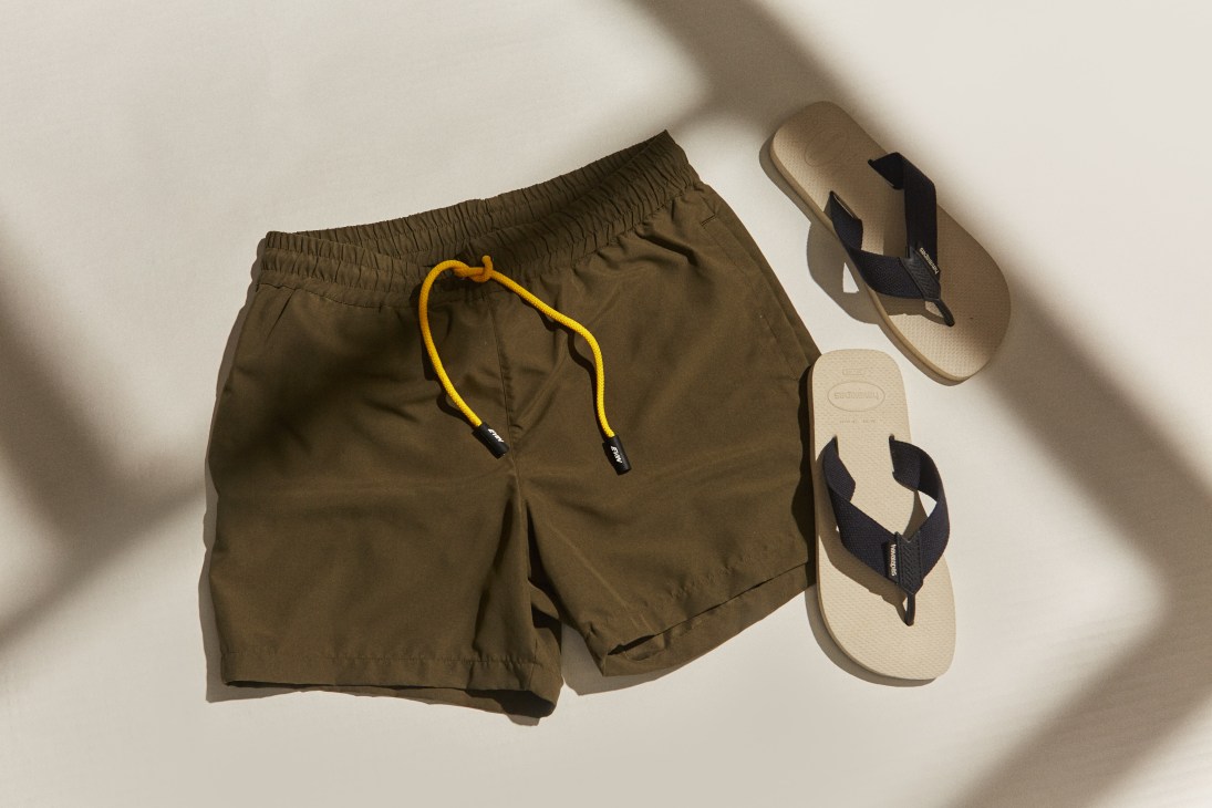 Olive-green swimming shorts with yellow drawstring and cream Havaianas flip flops on white background.