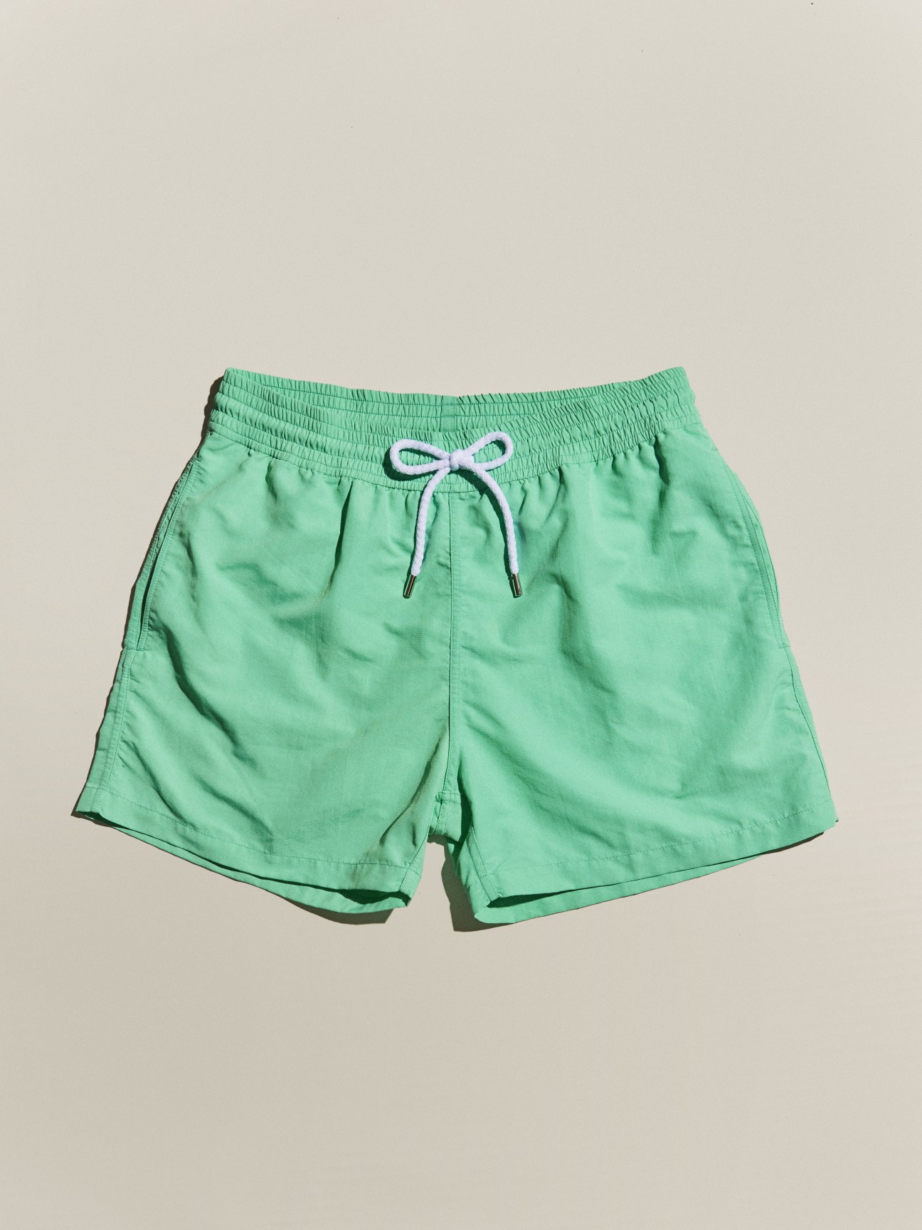 Green swimming shorts by Frescobol Carioca with drawstring waist on beige background.