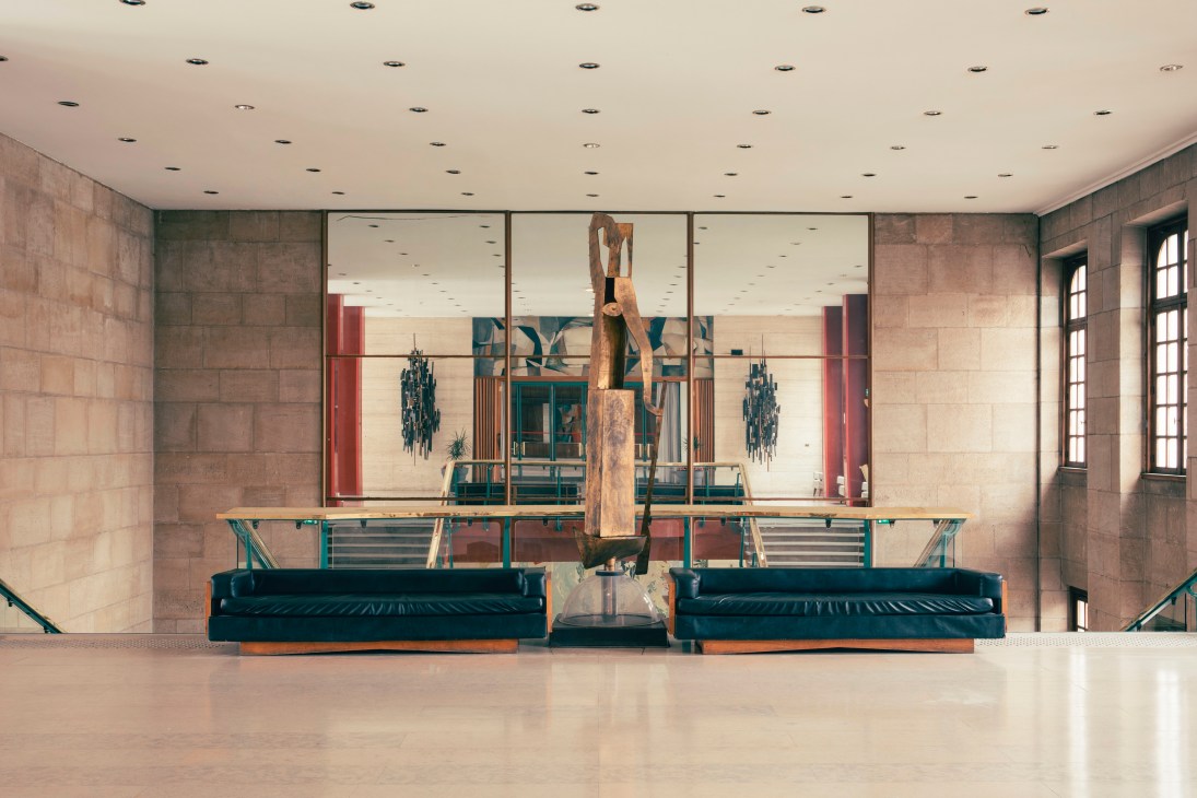 Modernist interior with black leather seating, glass table, and wooden sculpture in gallery space with stone walls