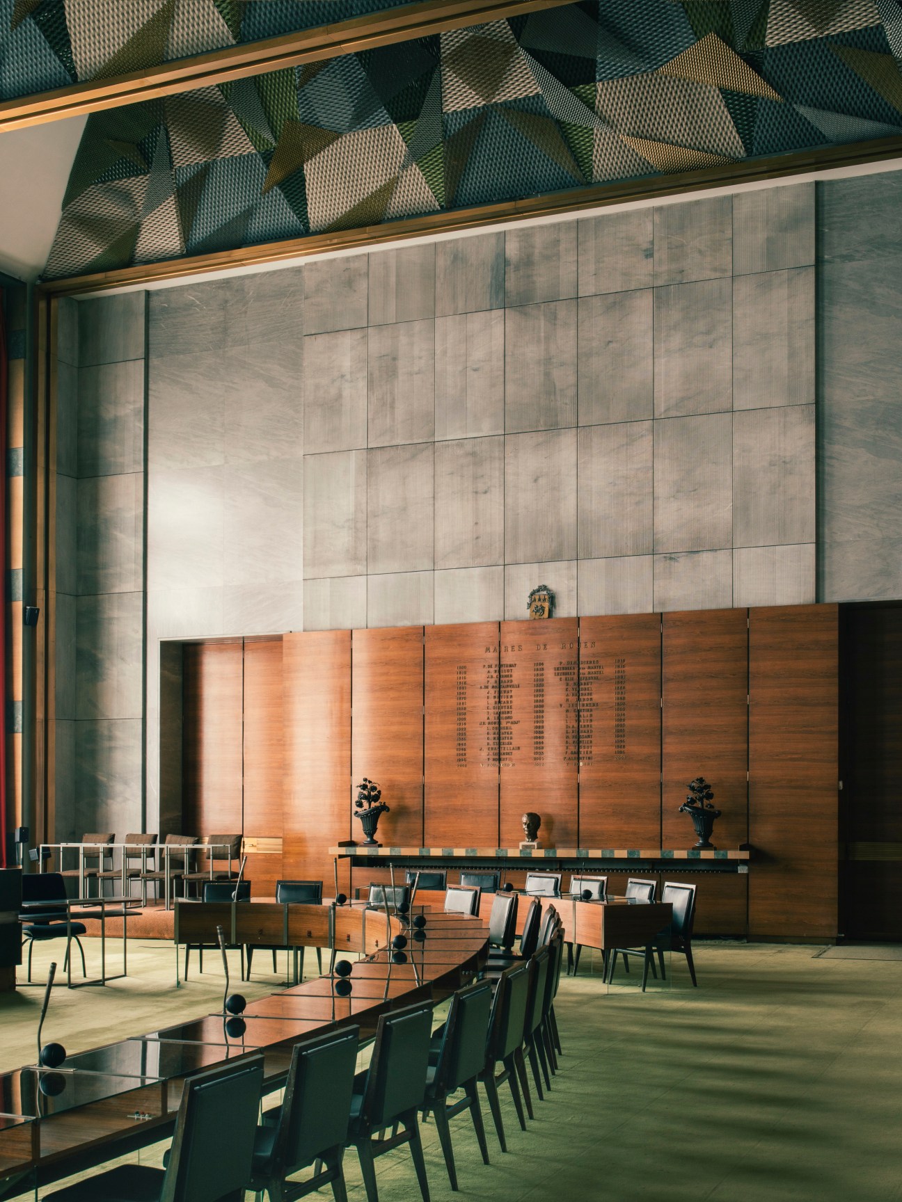 Modern council chamber interior with concrete walls, wooden panels, rows of dark chairs and decorative ceiling