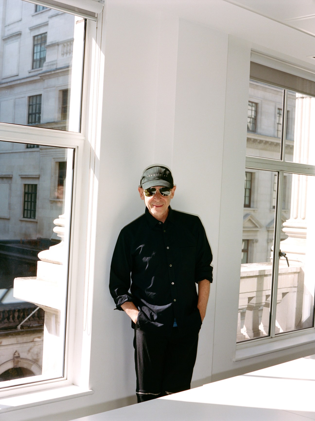 Adrian Joffe wearing a black cap, sunglasses, black shirt and trainers standing in a bright white office with large windows.