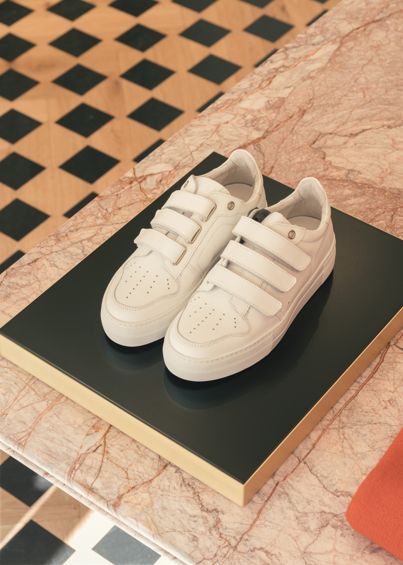 White leather low-top trainers with velcro straps displayed on a black box atop a marble surface with geometric tile pattern.