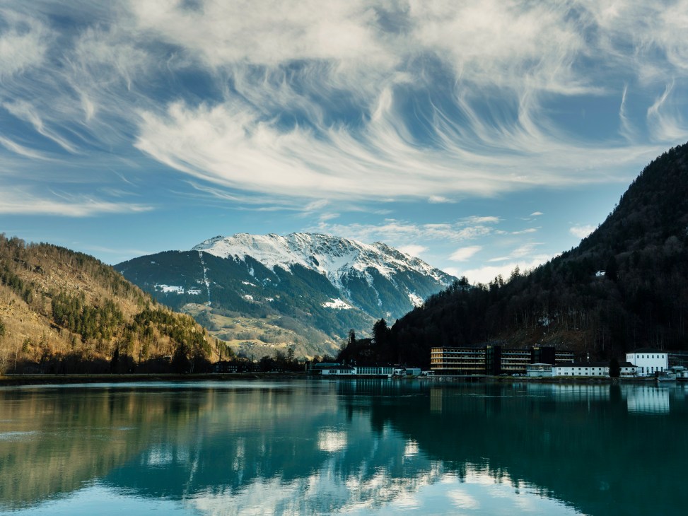 Vandans hydropower facility reflected in turquoise alpine lake surrounded by snow-capped mountains and forested slopes.