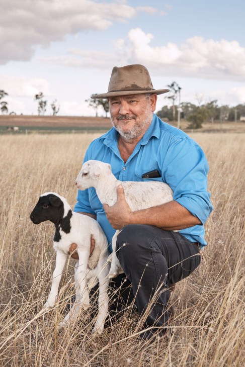 Local farmer Mark Williams holds two young goats in a rural field wearing a blue shirt and cowboy hat.