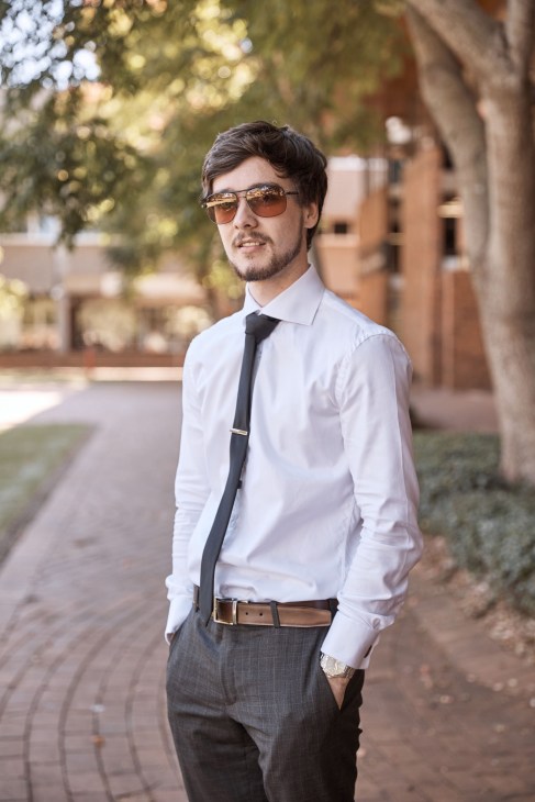 Man in white shirt, black tie and sunglasses standing on a tree-lined street.