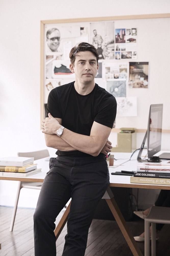 Designer David Caon sits on a desk in his studio, with inspiration board visible behind him.