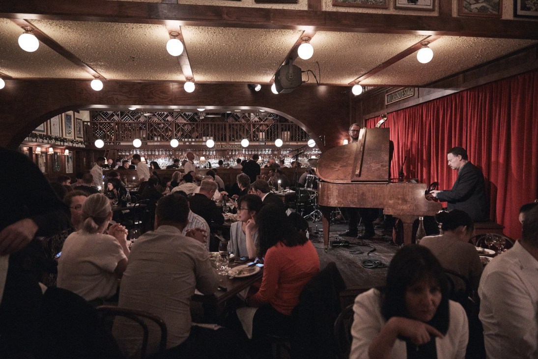Dimly lit dining room at Hubert restaurant in Sydney with red drapes, lit candles, and bow-tied waiters serving guests.