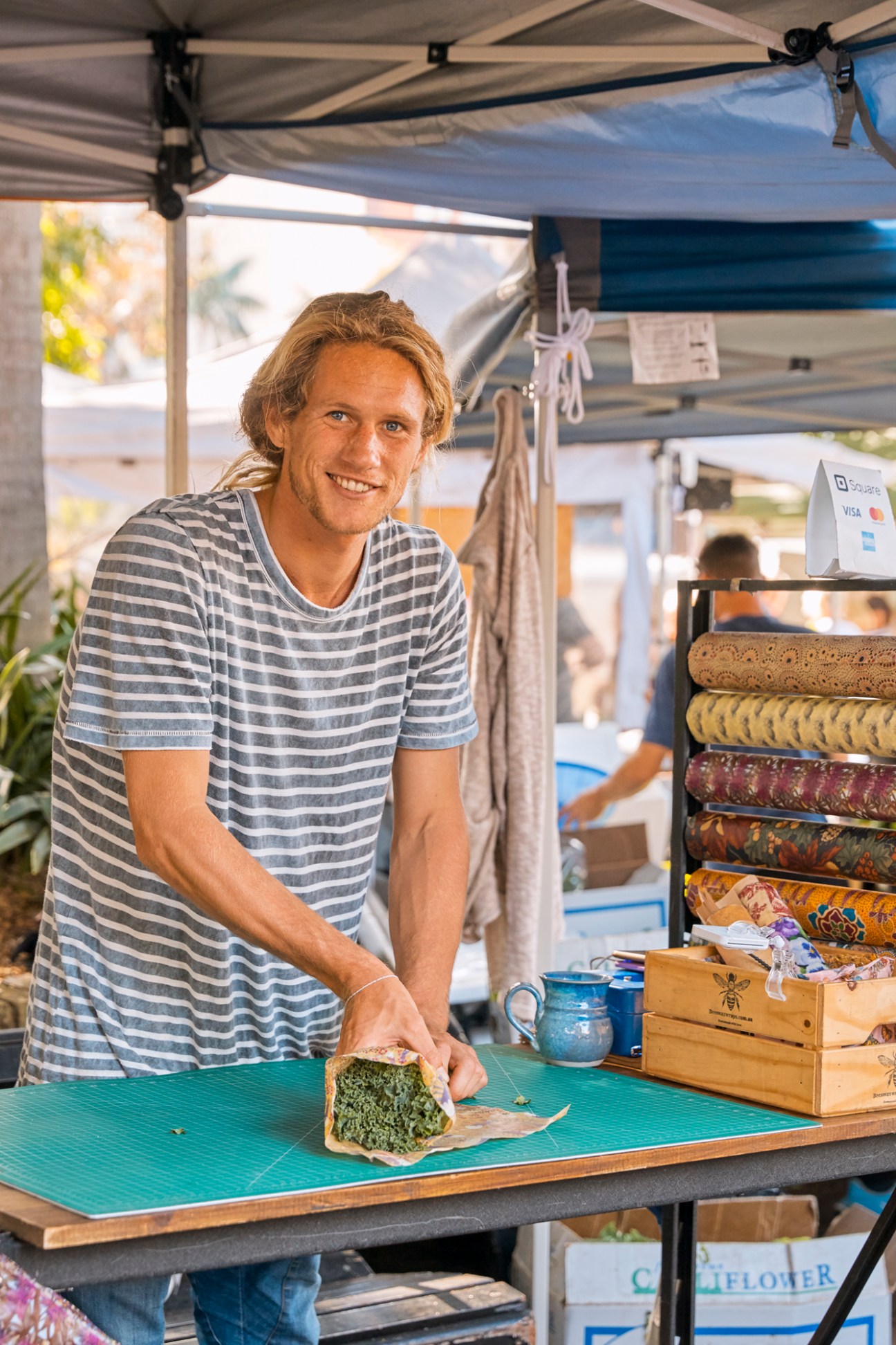 Man at market stall holding bread with beeswax wraps displayed on shelf behind him