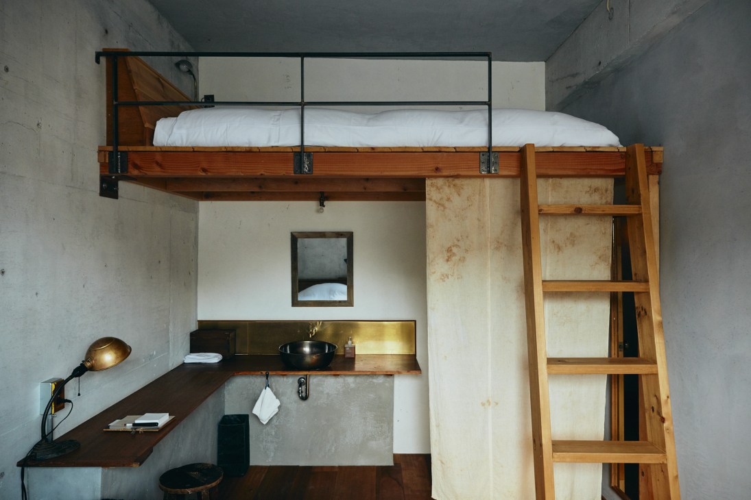 Minimalist hotel room with loft bed, wooden ladder, desk workspace, and concrete walls.