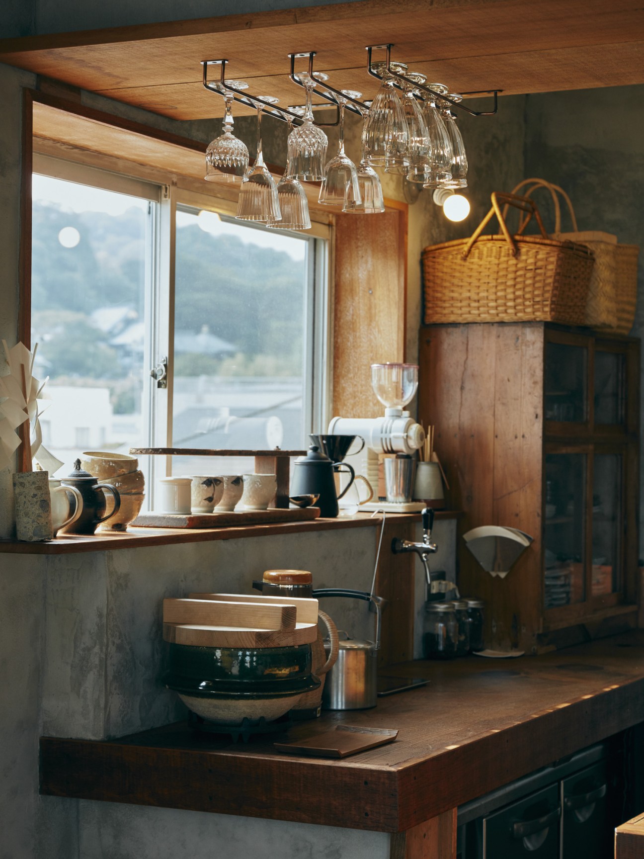 Coffee bar with hanging glassware, ceramic vessels, and woven baskets overlooking mountain landscape.