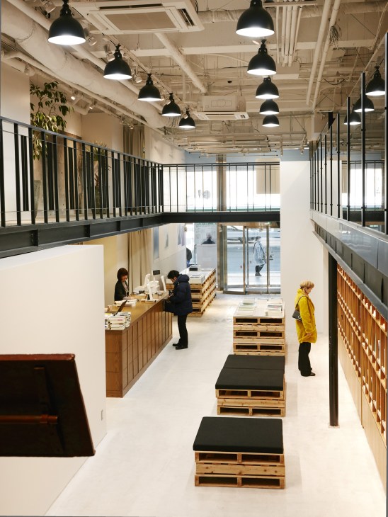 Interior of Bunkitsu bookshop showing mezzanine level with black pendant lights, wooden pallet seating, and customers browsing books.