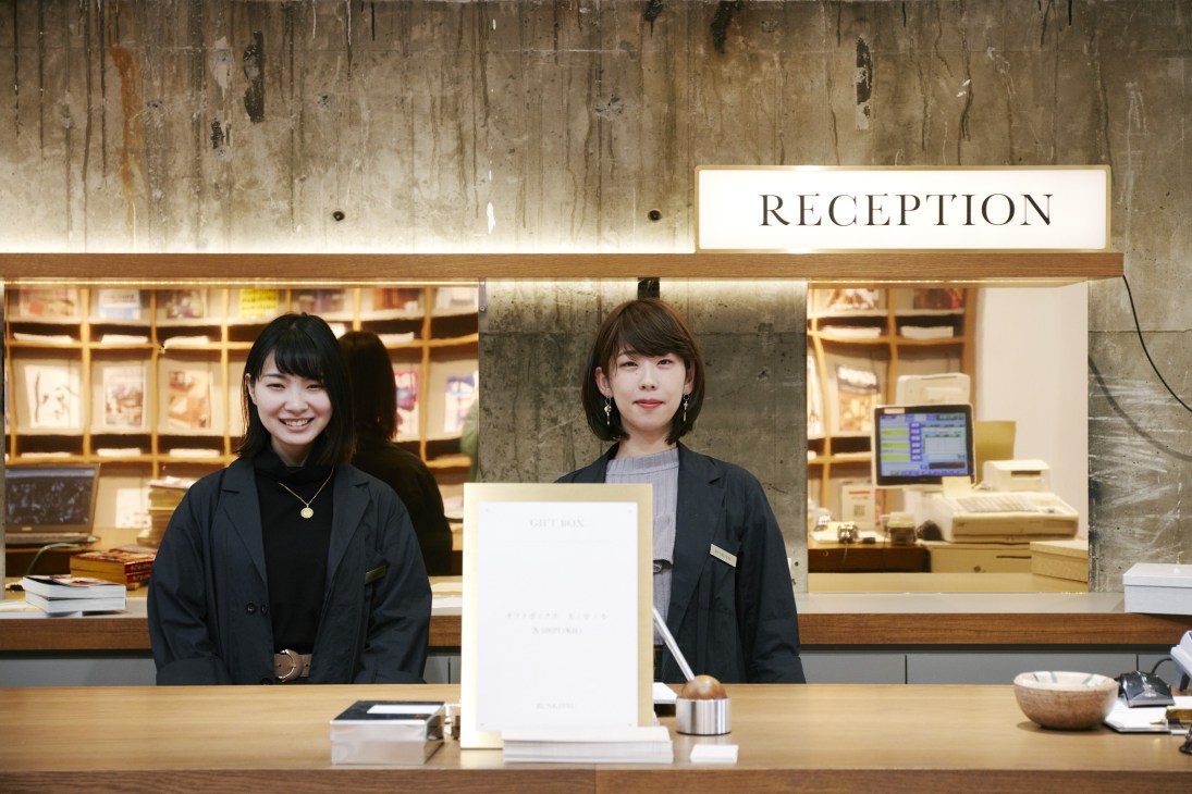 Two staff members at Bunkitsu's reception desk with illuminated bookshelves behind them.