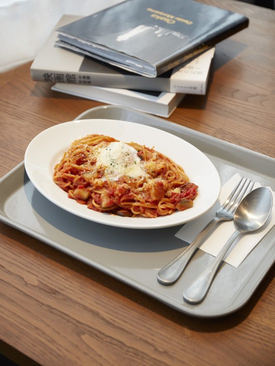 A plate of pasta with tomato sauce and cheese sits on a white tray with cutlery, beside stacked books on a wooden table.