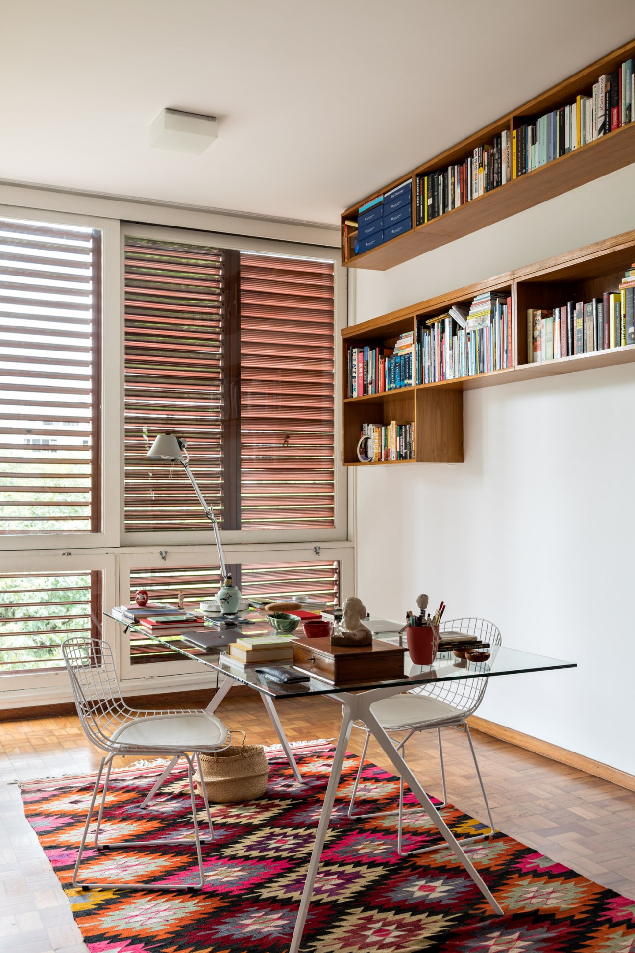 A modern study with wooden wall shelves, colorful horizontal window shutters, a glass desk, and wire chairs on a patterned rug.