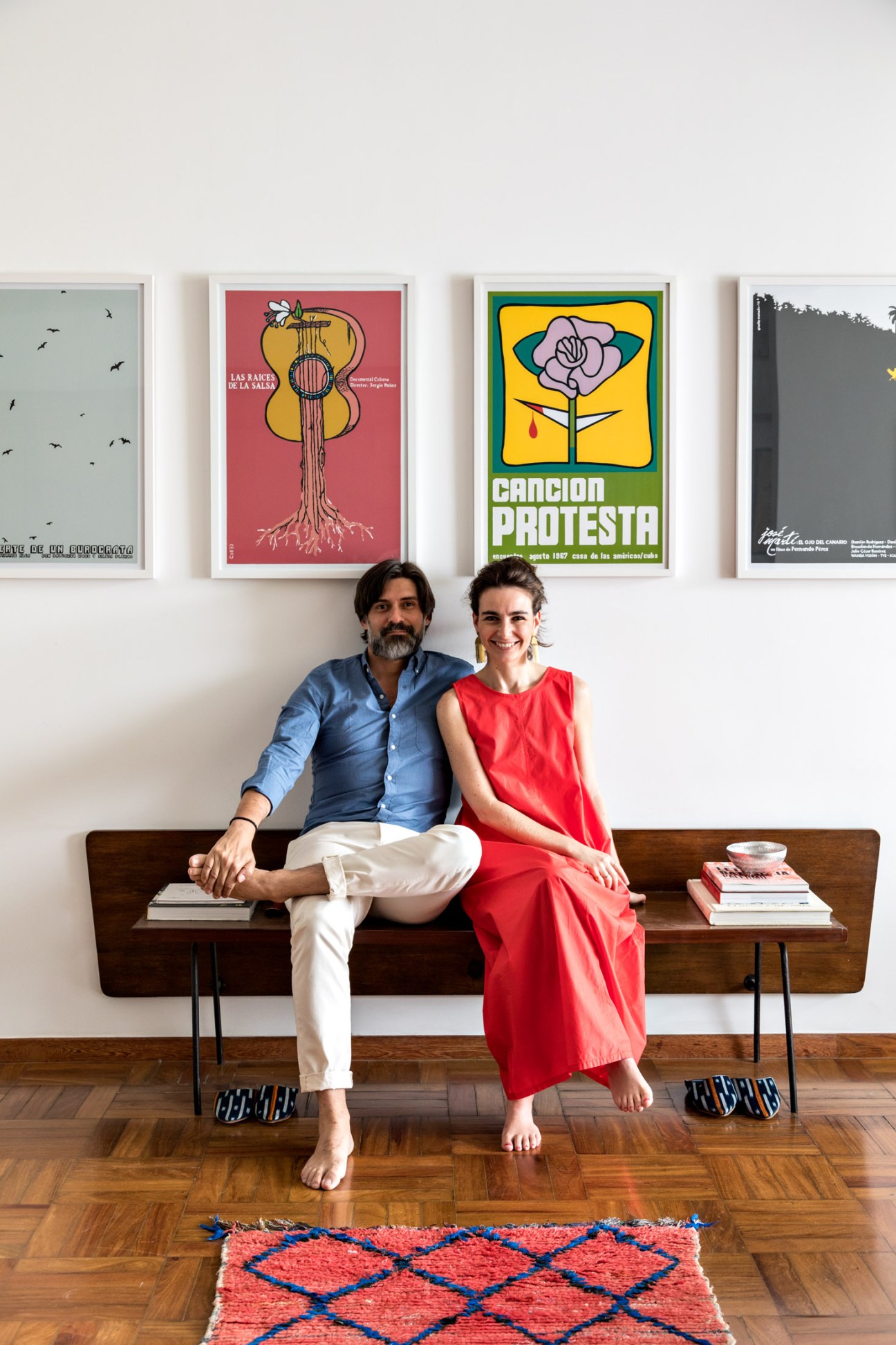 Andres Schipani and Alejandra Mejia sit on a wooden bench beneath vintage posters in their Lausanne apartment.
