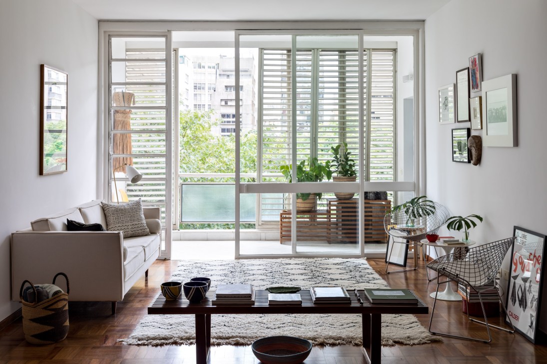 Modernist living room with large glass doors opening to a balcony with horizontal shutters, neutral sofa, dark coffee table, potted plants, and framed artwork on walls.