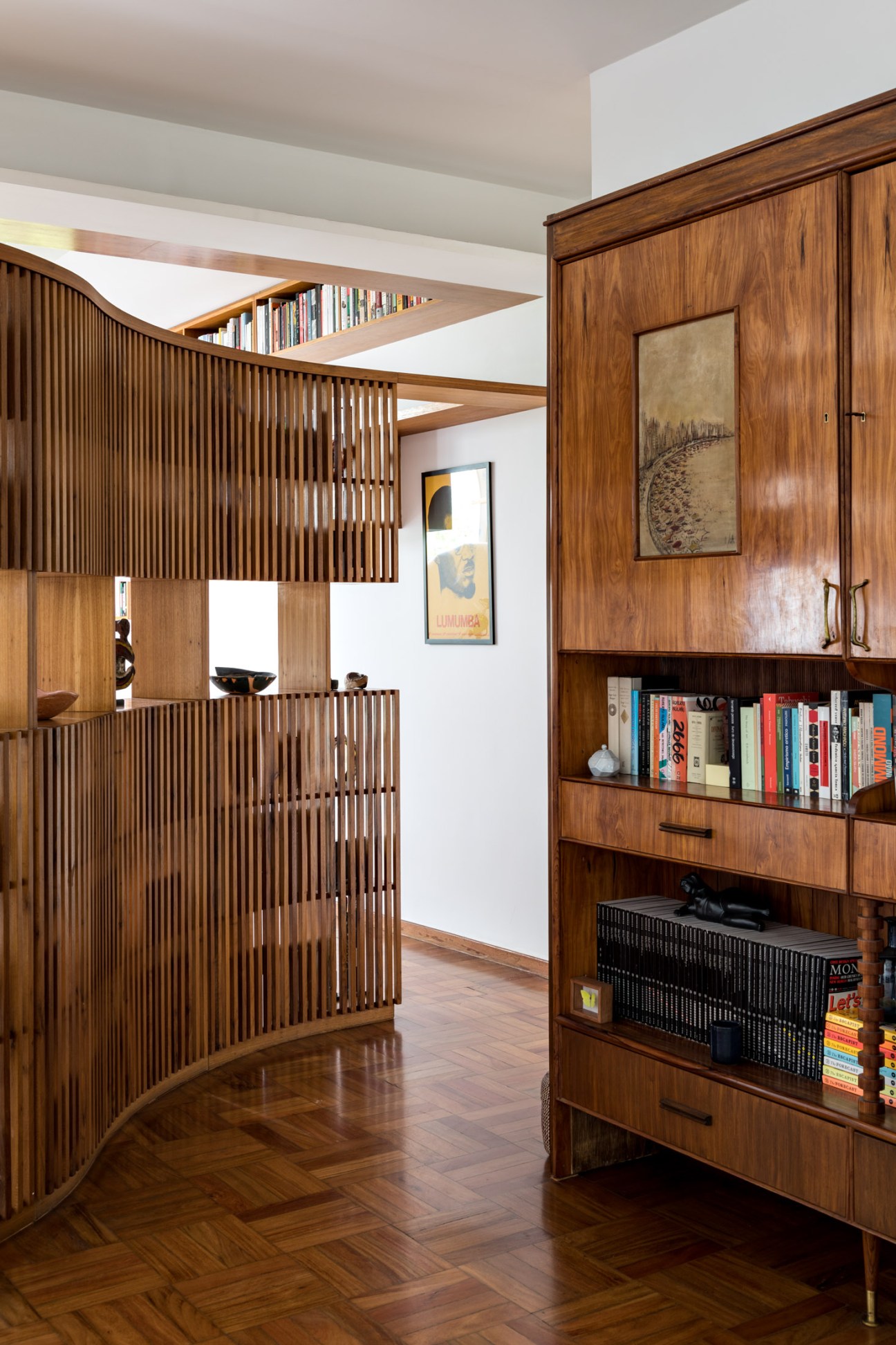 Curved wooden bookshelf with vertical slats filled with books, next to wooden cabinet with shelves in modernist apartment.