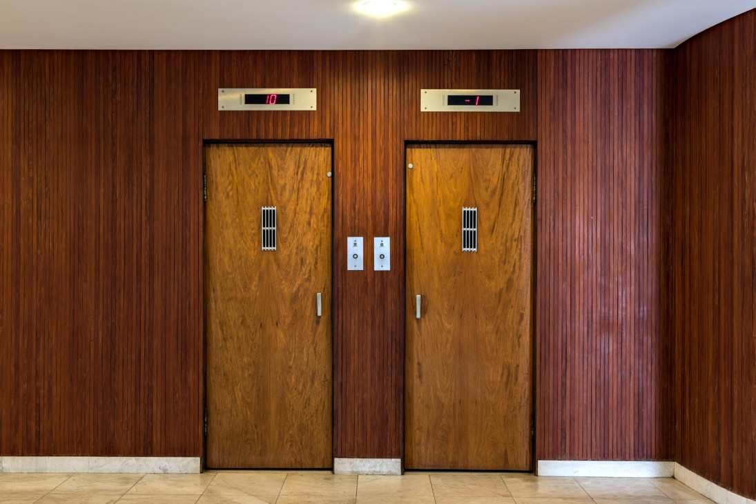 Two wooden elevator doors with floor indicators and call buttons in a modernist building hallway with vertical wood paneling.