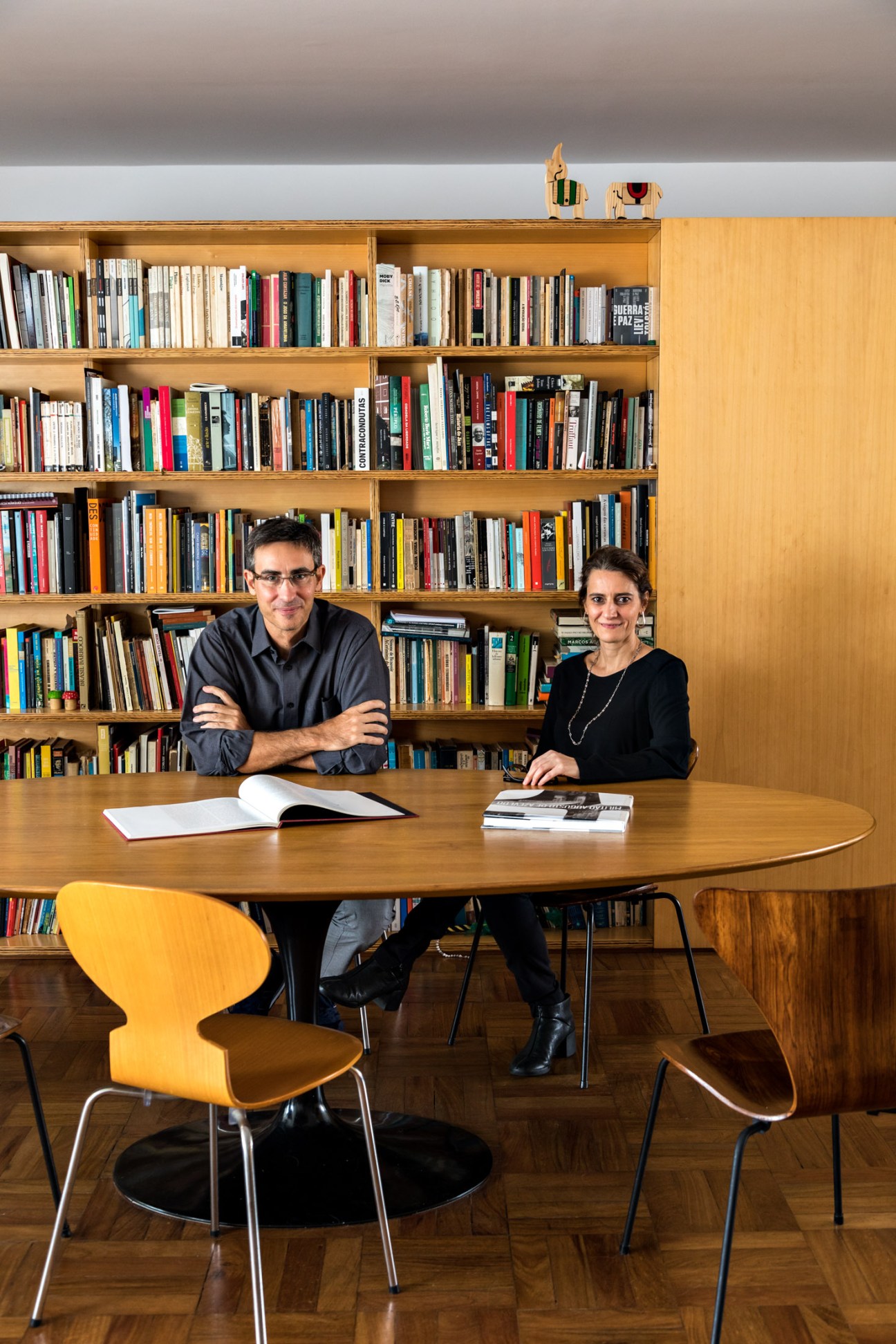 Two people seated at a wooden table in front of bookshelves in a modernist apartment.