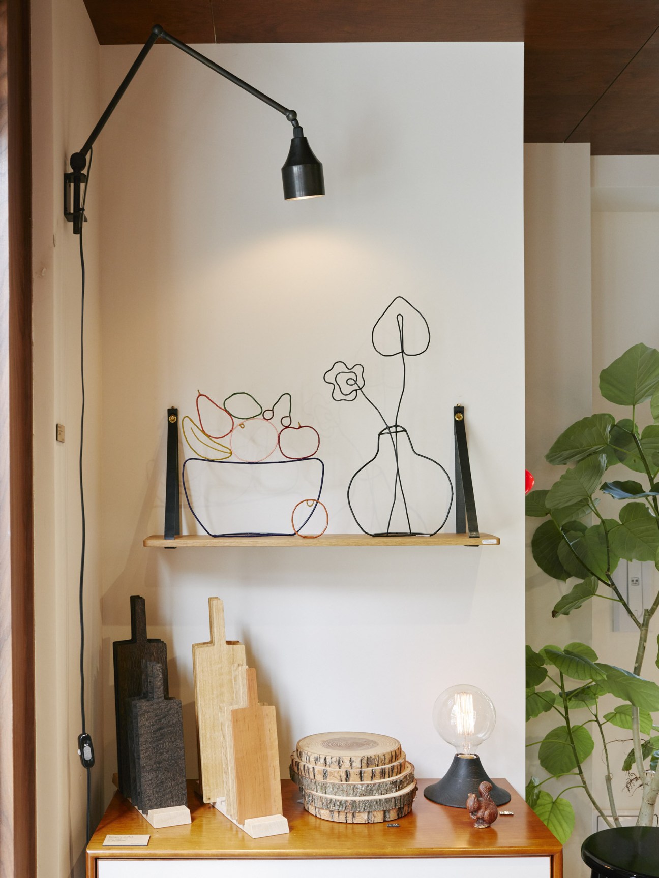 Bullpen furniture shop interior with white wall shelf displaying wire-drawn plant and vase illustrations, wooden cutting boards, and decorative items below.