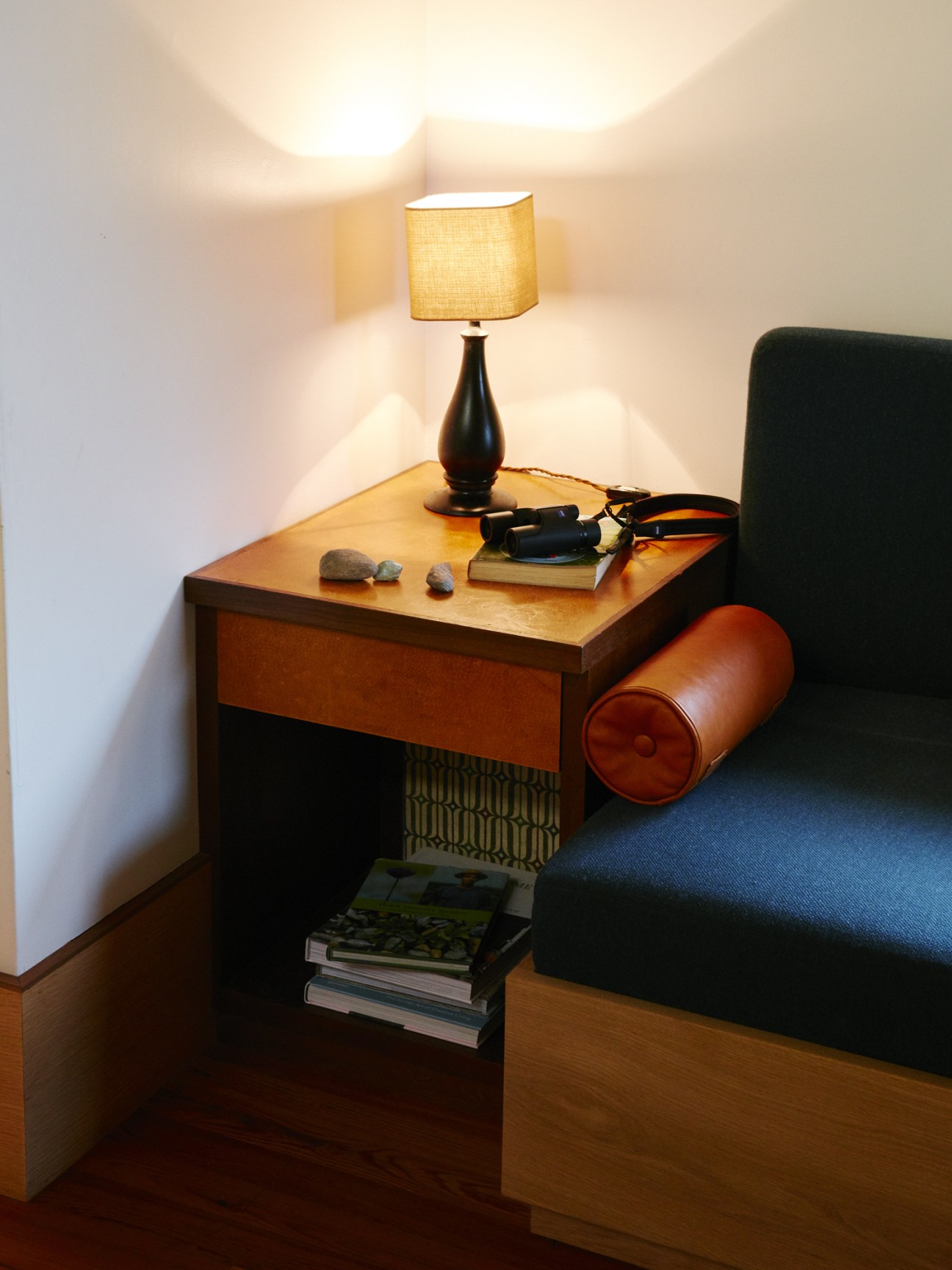 Wooden side table with a black table lamp, leather ottoman, and stacked books in a minimalist interior space.