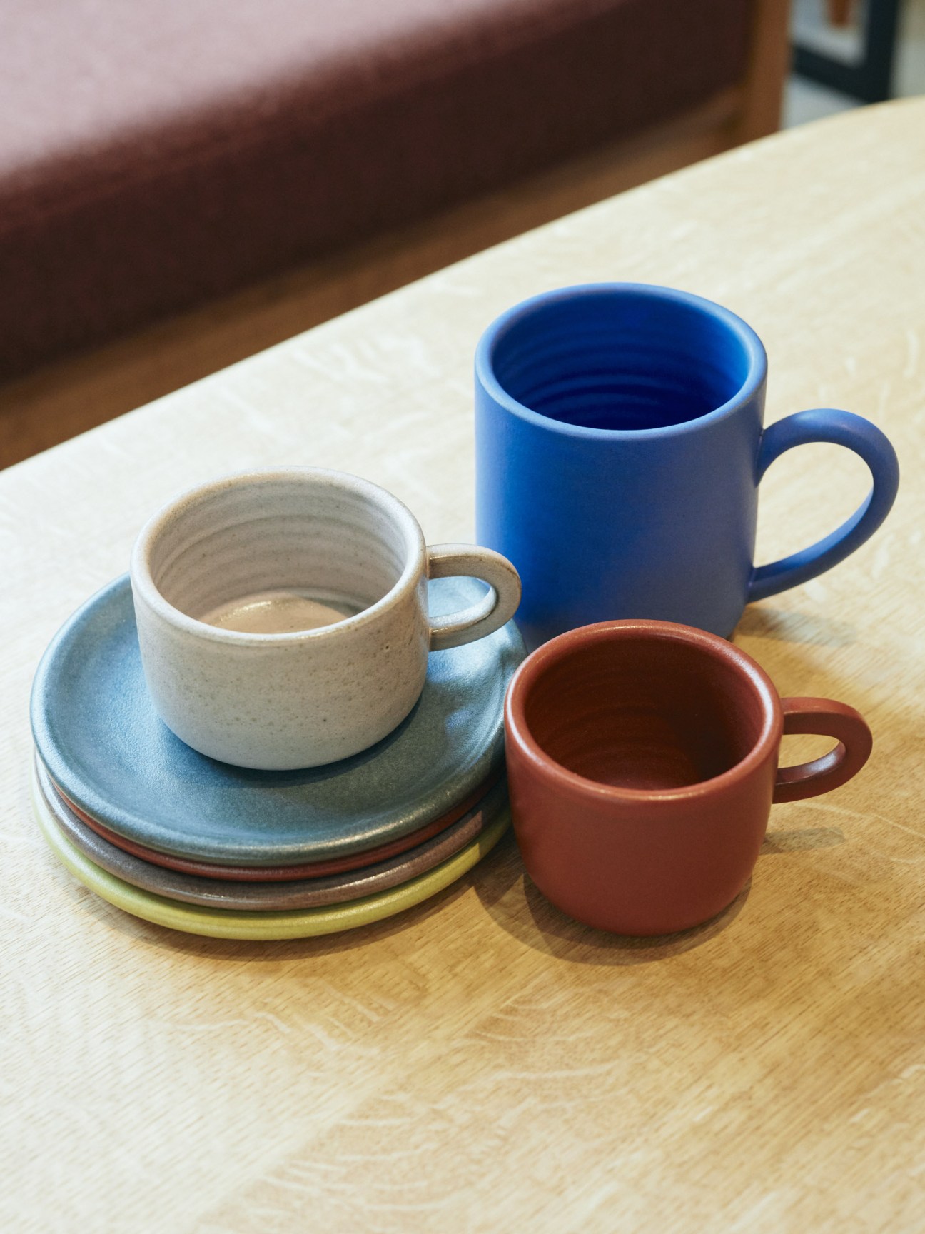 Handmade ceramic cups and saucers in cream, blue, and rust colors displayed on a wooden table.