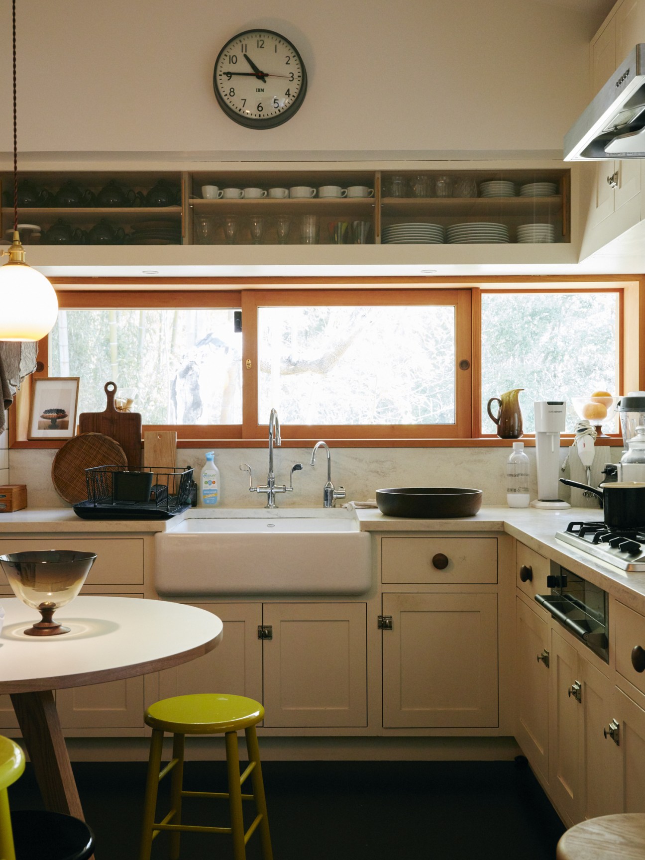 Bright kitchen with light wood cabinetry, white countertops, large windows, wall-mounted clock and open shelving above sink.