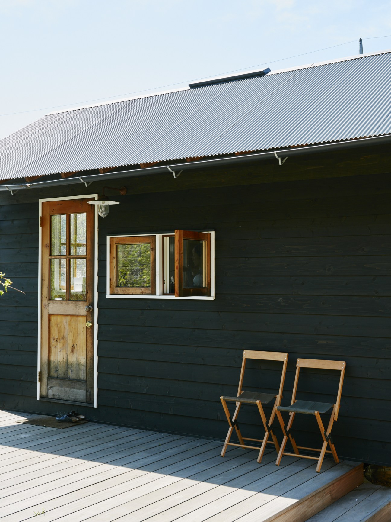 Kazuko Burioka's small baking hut with dark exterior walls, wooden door, windows, and two wooden chairs on a wooden deck.