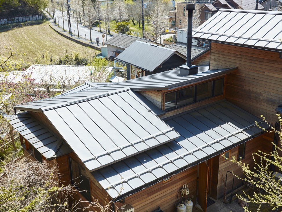 Aerial view of a renovated Japanese house with red cedar siding and metal roofs surrounded by green landscape in Tokyo's Machida city.
