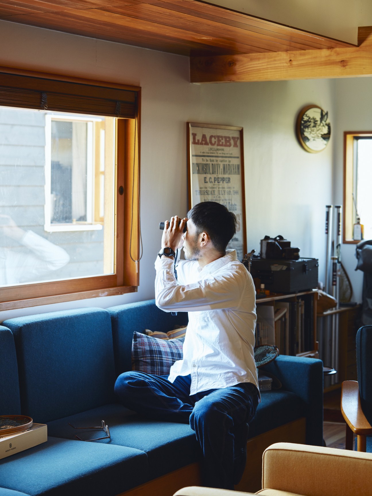 Man with binoculars sitting on blue sofa by window birdwatching.