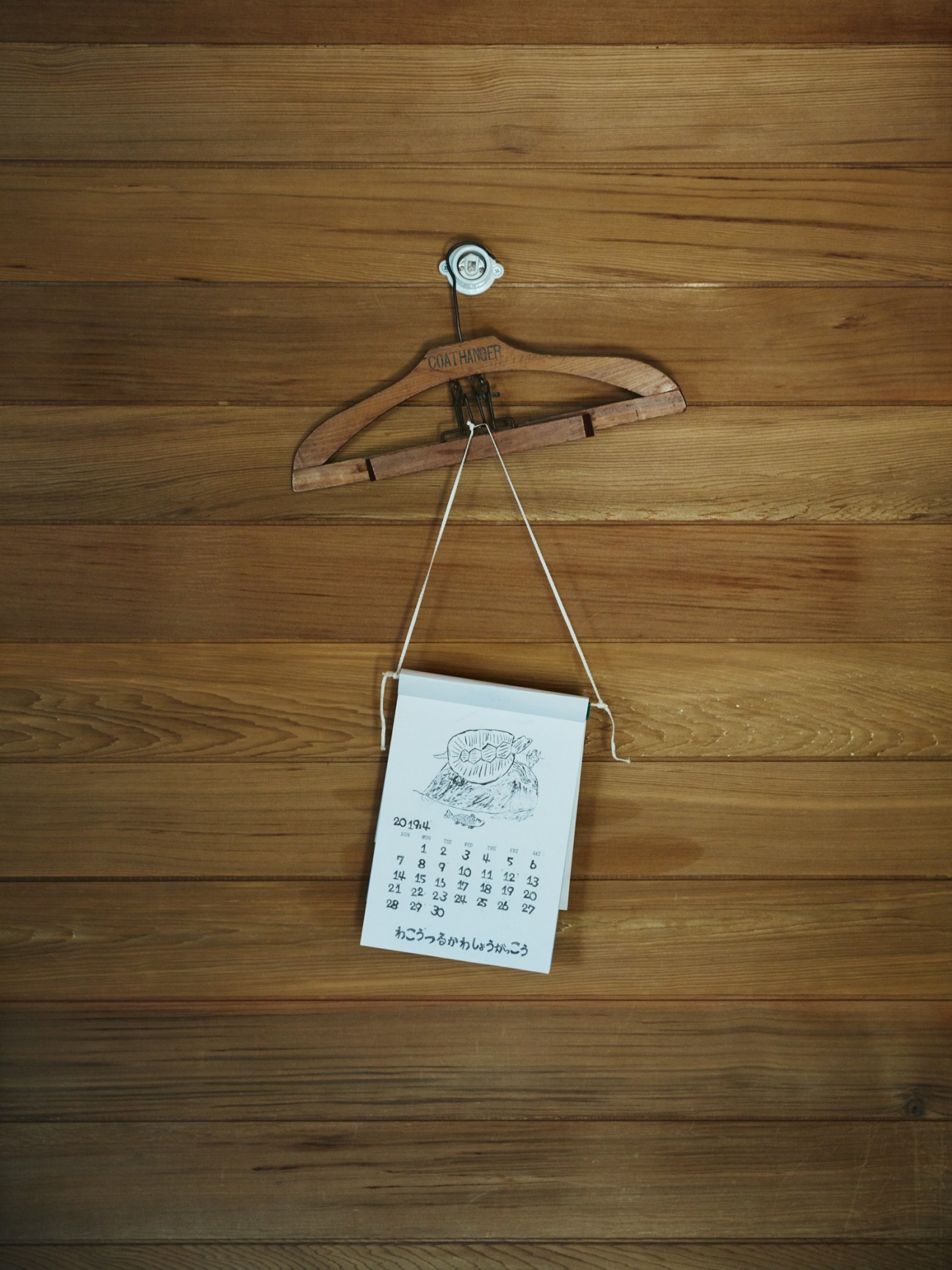 A wooden coat hanger holding a calendar with Japanese text mounted on aged red cedar wall paneling.