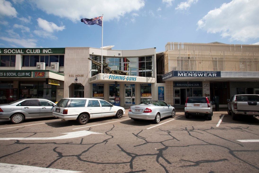 Darwin city centre storefronts including a social club, fishing-guns shop, and menswear store with parked cars in front.
