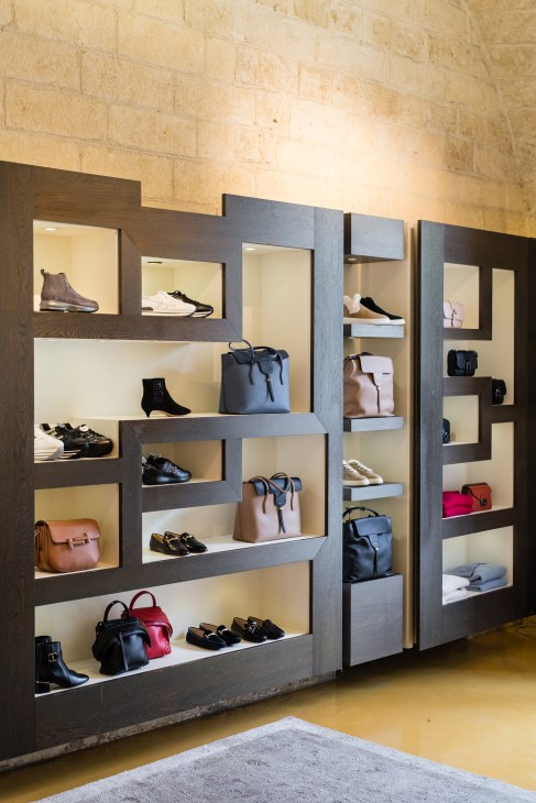 Luxury boutique display with dark wooden shelving units containing shoes, handbags, and accessories against a cream stone wall.