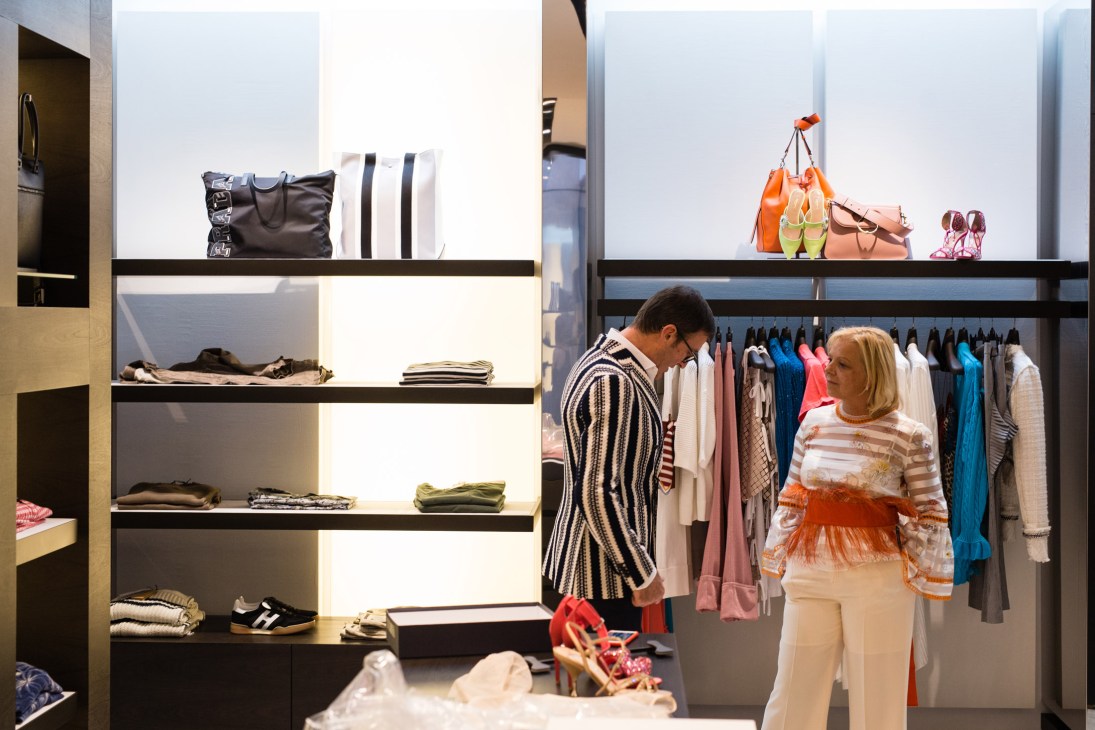 Modern boutique interior with illuminated shelving displaying folded clothing, handbags, and shoes, with two customers browsing.