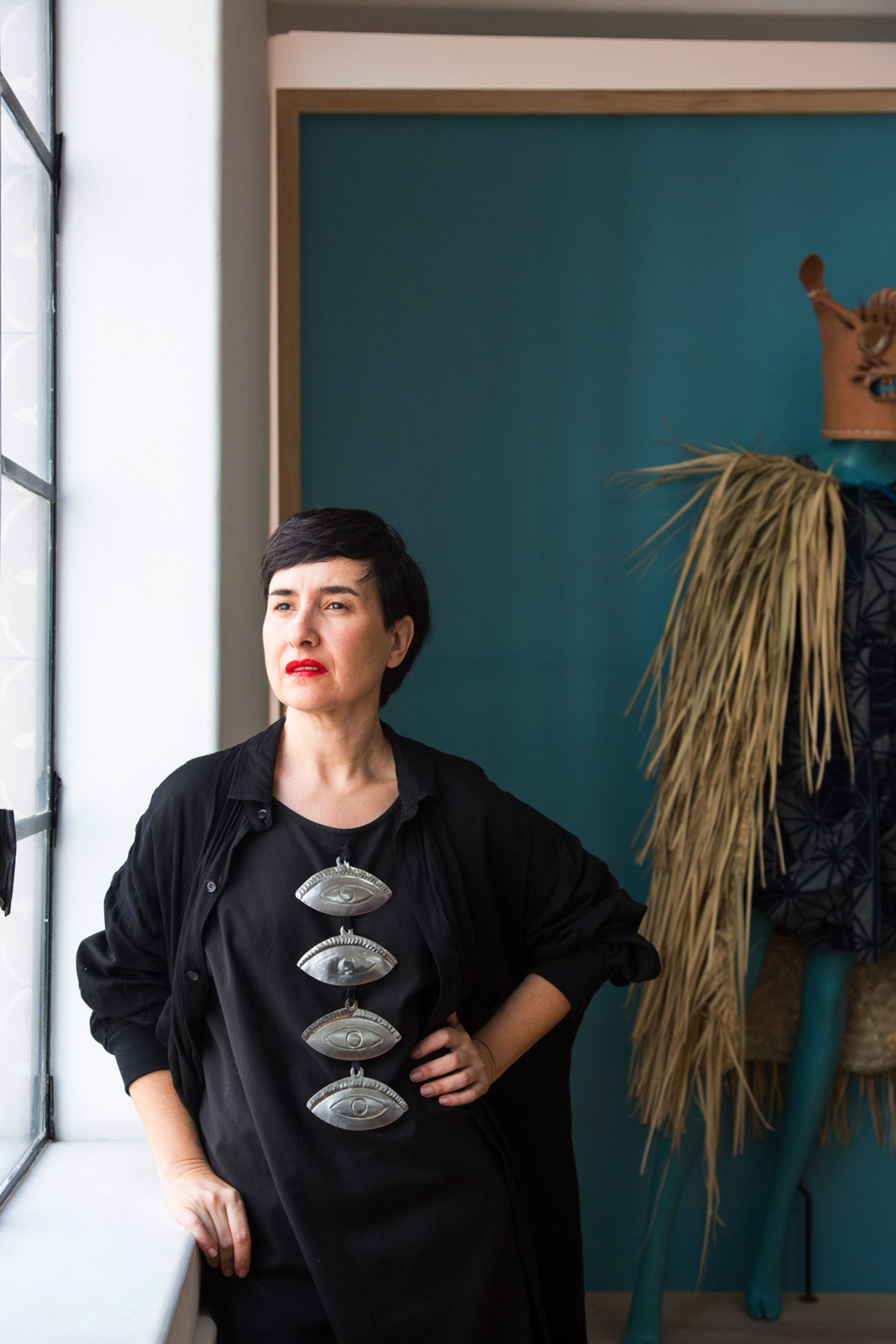Designer Carla Fernández wearing a black jacket and eye-motif shirt beside clothing with fringed details against a teal wall.