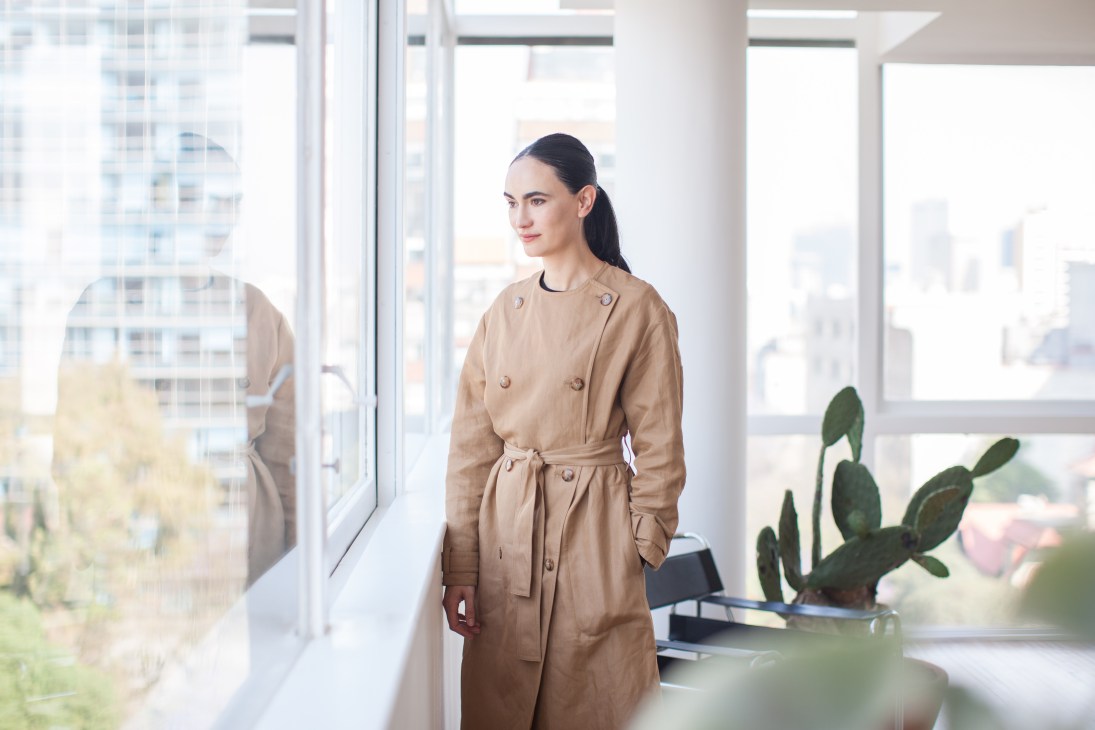 Frida Escobedo stands by a window in a modern office, wearing a beige trench coat.