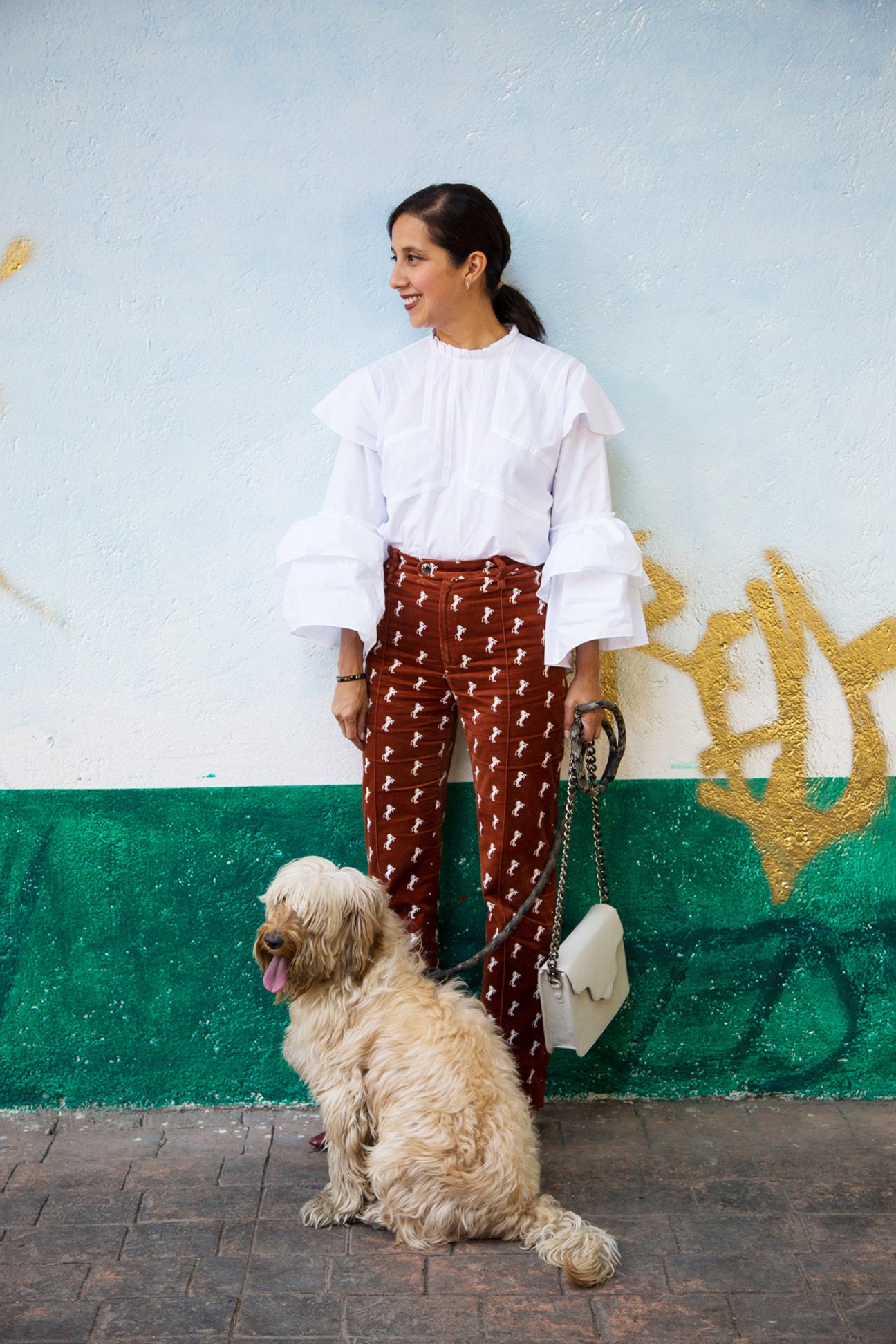 Karla Martínez de Salas wearing a white ruffled blouse and patterned rust-brown trousers against a colorful wall.