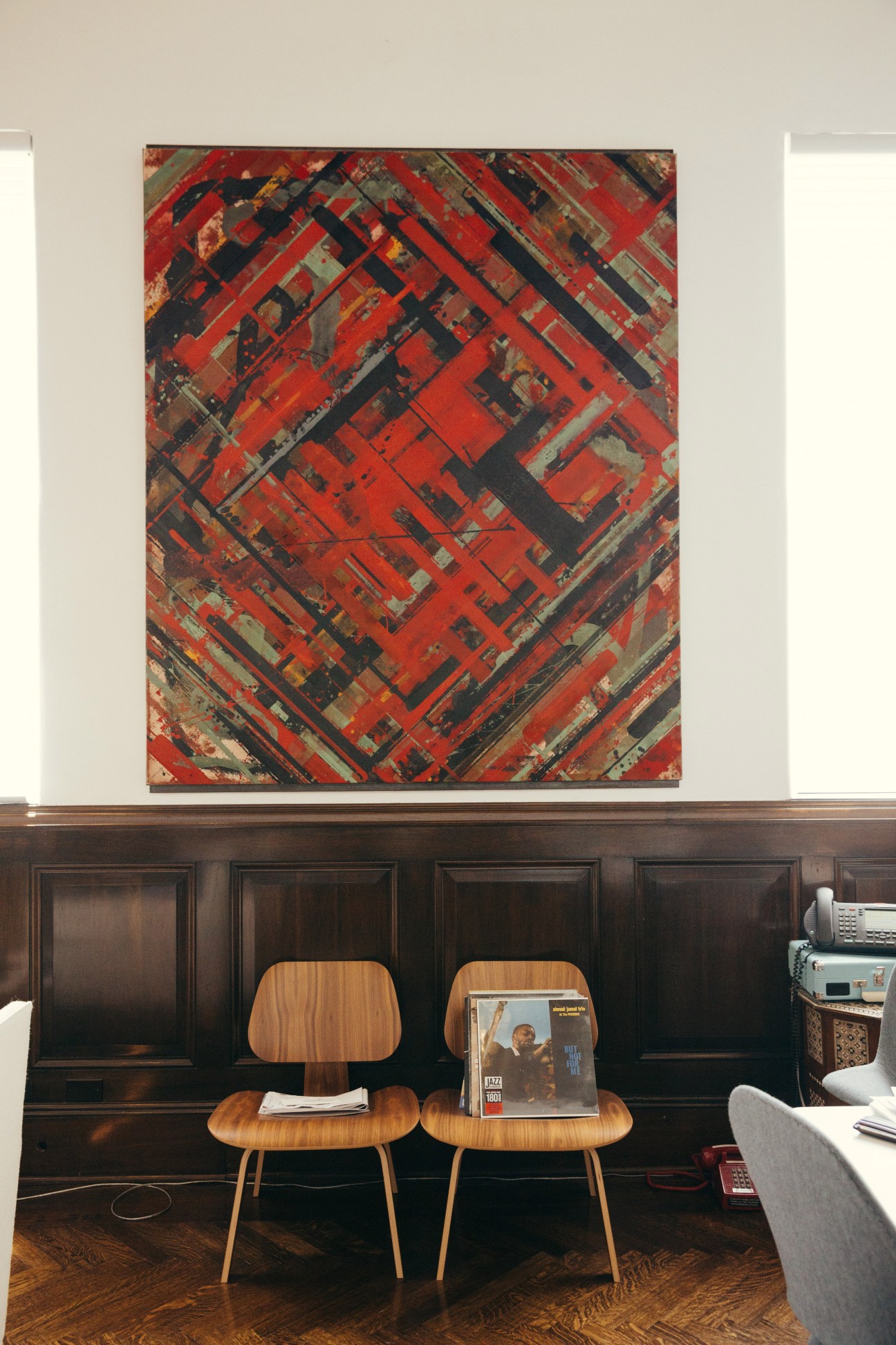 Abstract artwork with red, black and tan geometric patterns hanging on a white wall above wooden chairs.