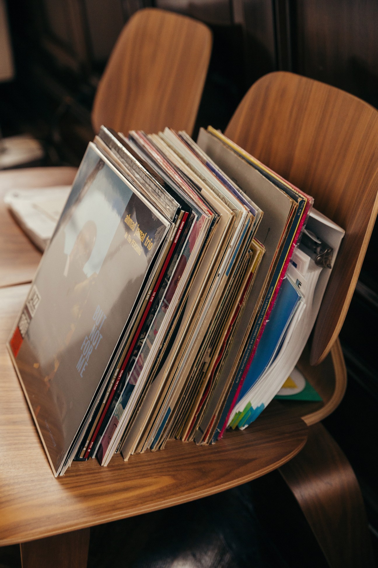 Vinyl records stored in a wooden holder on a table with wooden chairs visible in the background.