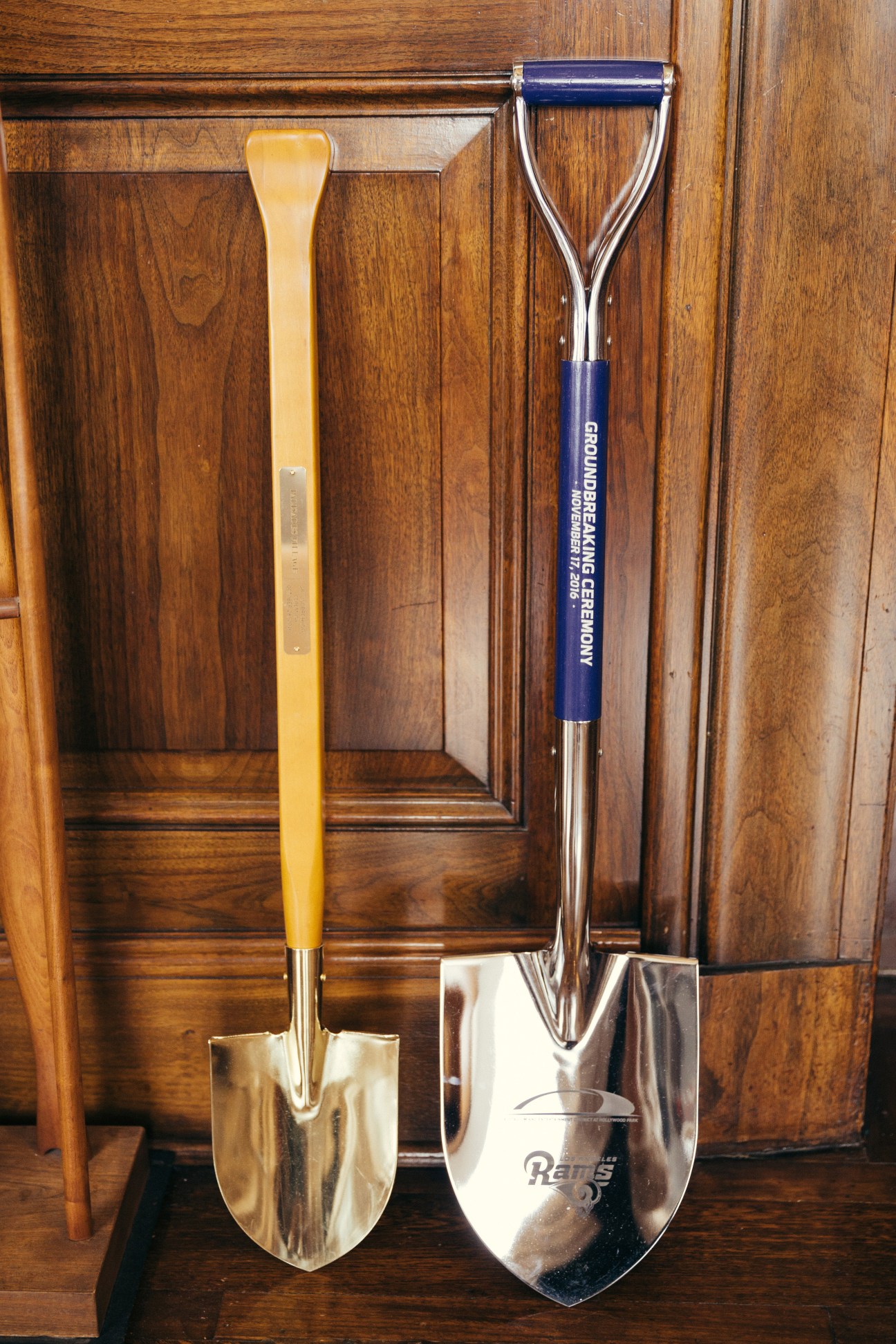 Two ceremonial shovels with wooden and blue handles leaning against wooden paneling in a city hall office.