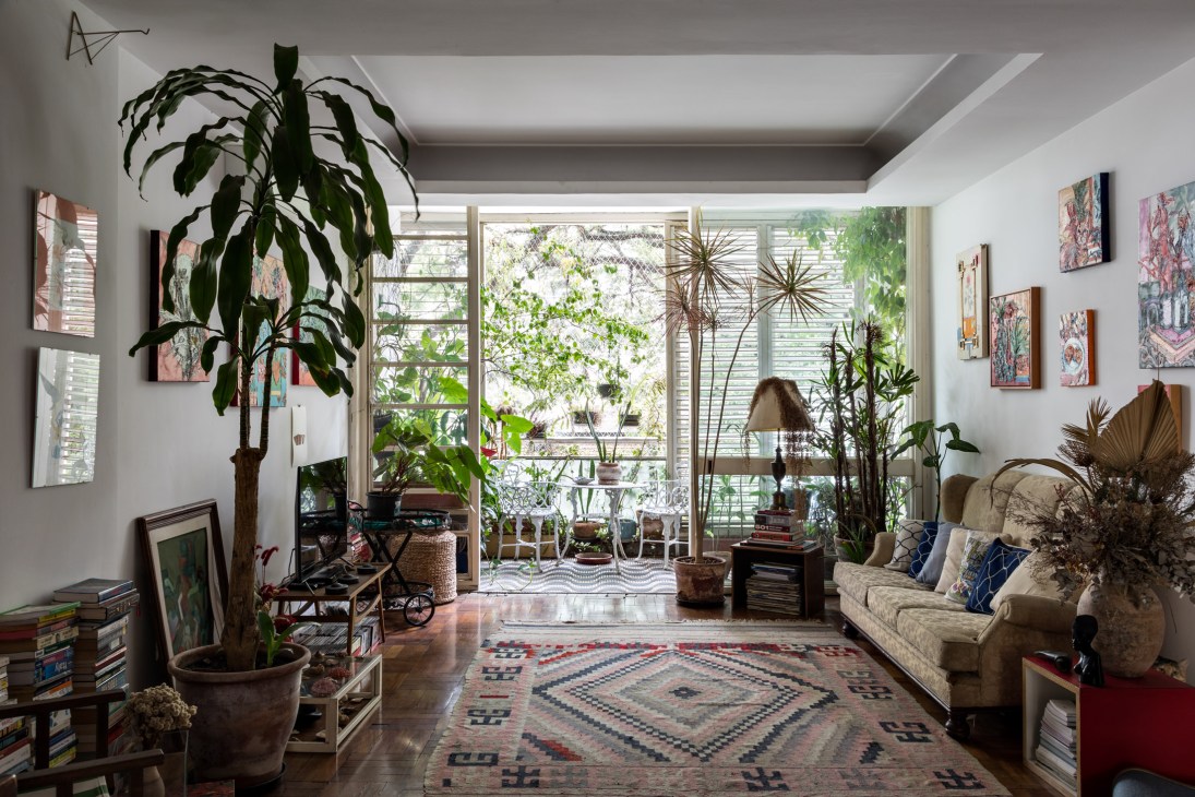 A bright living room with high ceilings, lush potted plants, and views of a balcony through floor-to-ceiling windows with adjustable shutters.