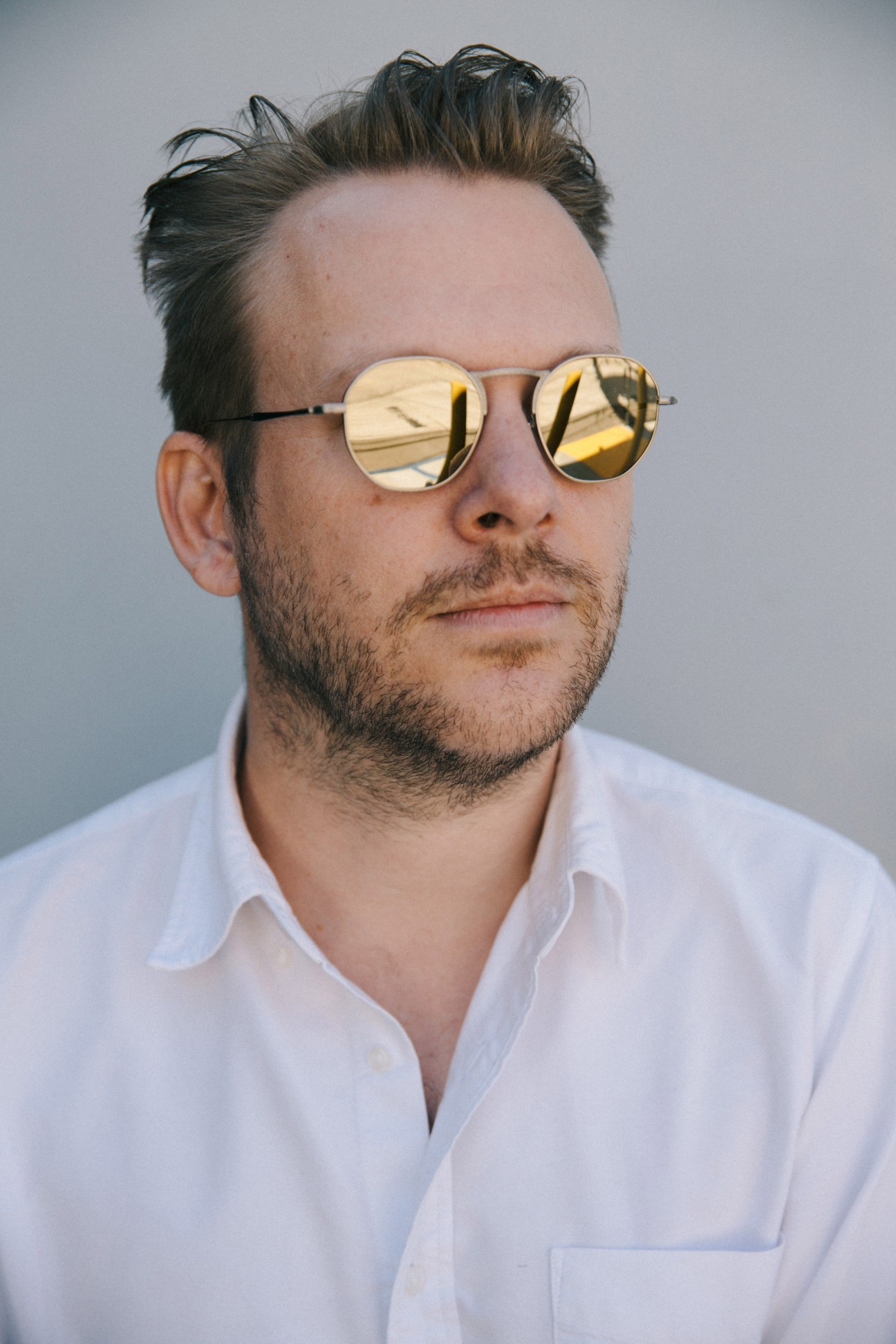 Man wearing round gold-mirrored sunglasses and white shirt against neutral background